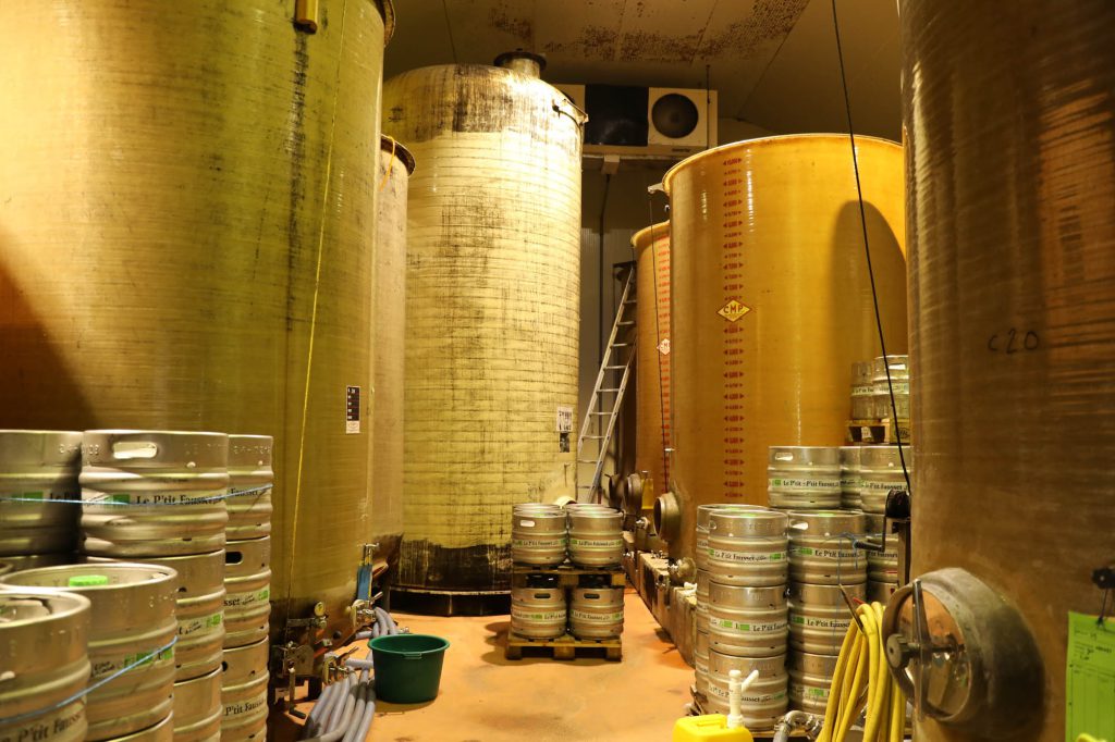 Cellier avec cuves jaunes de fermentation et fûts inox Le P'tit Fausset empilés